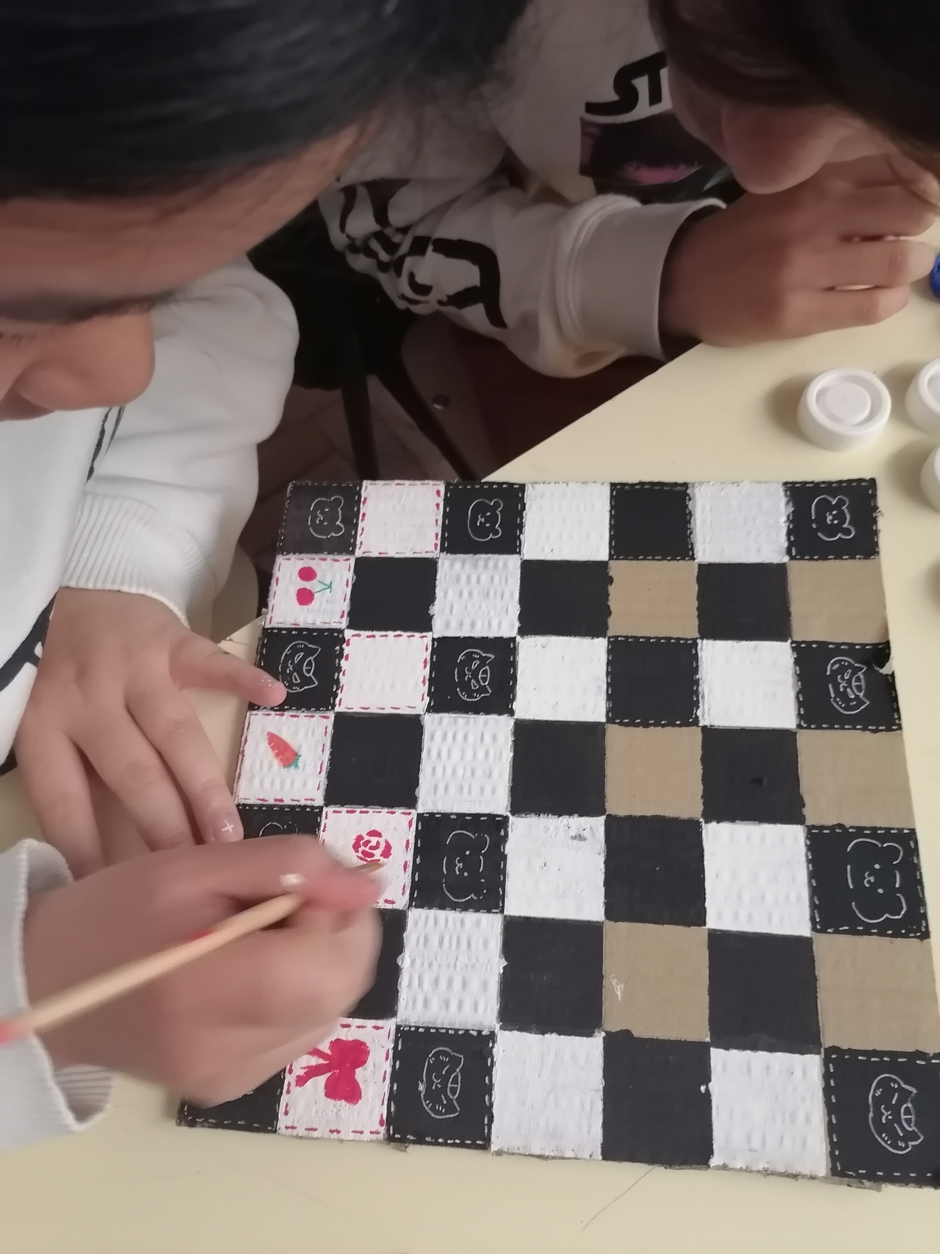 Pintura pormenorizada das quadrículas do tabuleiro de jogo; demonstração de sentido estético e minucia acompanhados do olhar maravilhado dos colegas de grupo